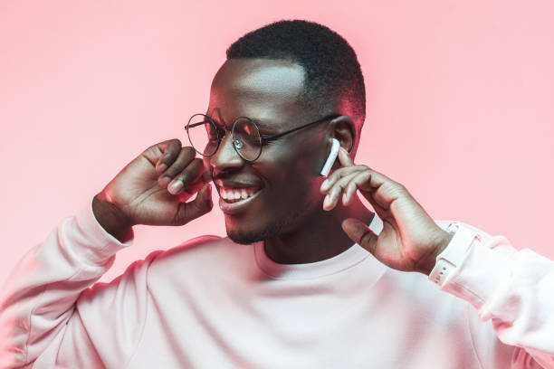 portrait en studio de heureux jeune homme africain, écouter de la musique avec des écouteurs sans fil isolés sur fond rose - men fashion model cool glasses photos et images de collection