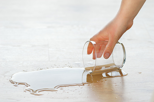 cropped shot of a woman hand picking up a drinking glass of water spilled over the laminated wooden floor with a puddle of water
