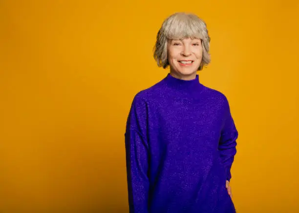 Portrait of a mature woman in front of a yellow background. She is wearing a purple sweater.