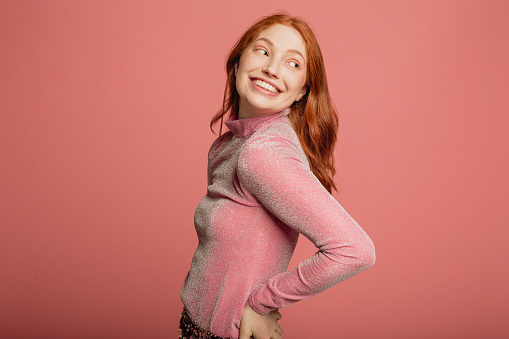 Close-up portrait of a young redhead woman dressed in a pink sparkly top standing in front of a pink background.