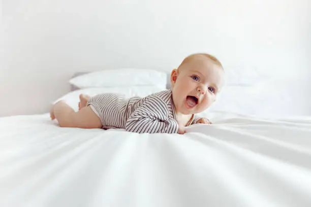 Side view of adorable little baby lying on white soft blanket on bed smiling excitedly in daylight