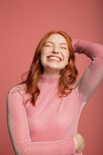 Close-up portrait of a young redhead woman dressed in a pink sparkly top standing in front of a pink background.