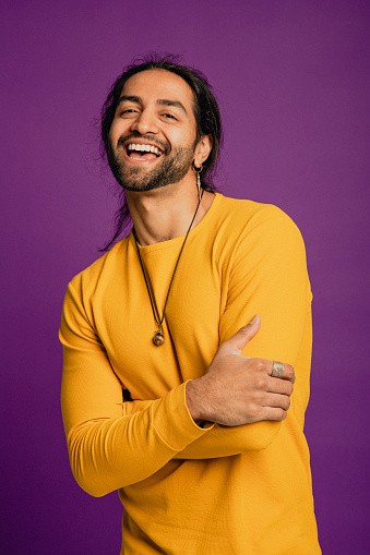 Portrait of an Asian man laughing, he is standing in front of a purple background and wearing a yellow sweater.