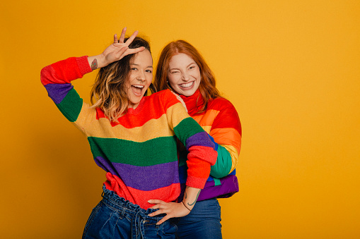 Portrait of a young redhead woman and a young mixed race woman both dressed in striped rainbow tops, standing in front of a yellow background.