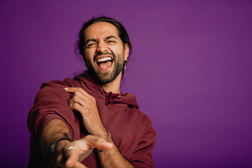 Portrait of a handsome Asian man wearing a purple hoodie yelling and pointing towards the camera in front of a purple background.