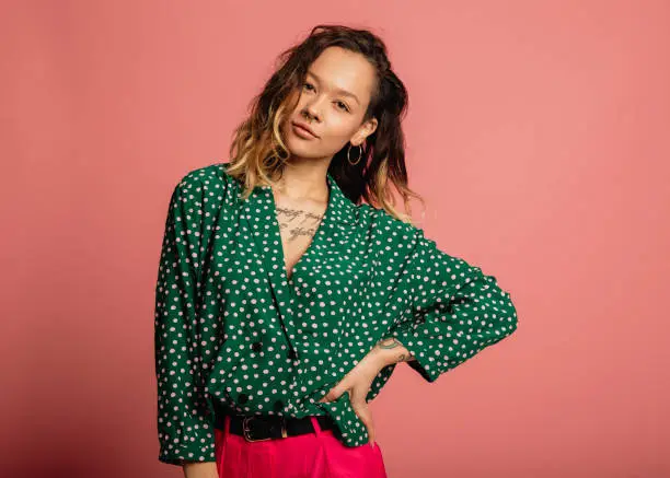Young woman dressed in a green polka dot shirt standing in front of a pink background.