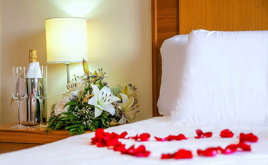 Horizontal perspective of a romantic setup with honeymoon bed with focus on the bouquet and champagne bottle and flute. White bedding, flowers, champagne flutes, concept for romance, honeymoon, or Valentine Day.