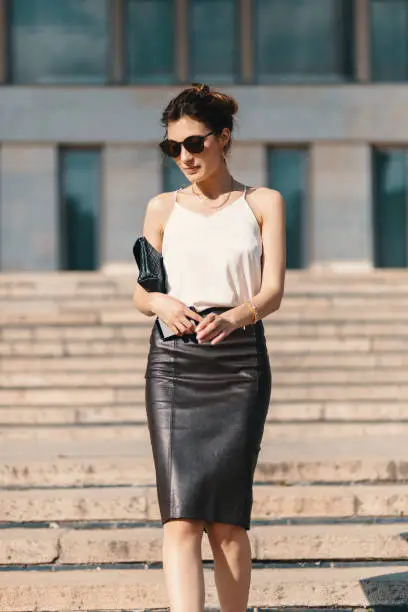 Fashionable brunette businesswoman in a brown leather pencil skirt and cream silk blouse, sunglasses walking in the street, on stairs. Fashion spring, summer photo. Long legs