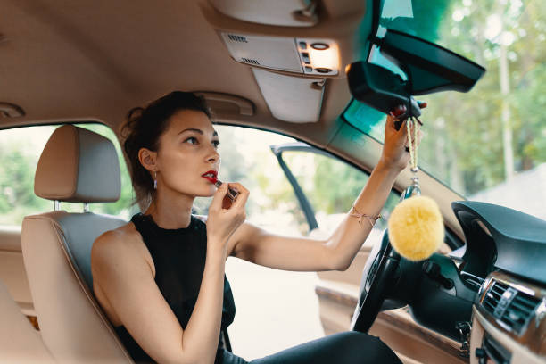 Young elegant woman looking in the car view mirror while applying makeup, lipstick on the lips