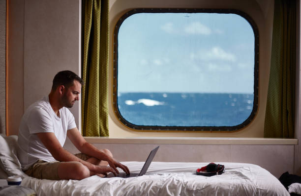 Working in a cabin while at sea A man sitting on a bed in a cabin on board a cruise ship. He is sitting on a bed, working at his computer, while out of the window the sea and sky can be seen. cruise ship people stock pictures, royalty-free photos & images