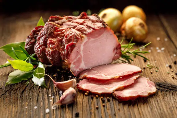 Photo of Sliced smoked gammon on a wooden  table with addition of fresh  herbs and aromatic spices.
