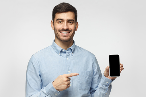 Handsome man in blue shirt isolated on gray background, presenting smartphone and pointing with finger at blank black screen with copyspace for ads