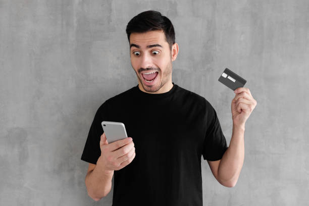istock Excited young handsome man standing isolated against gray textured wall background with mouth open, holding credit card, using mobile phone 1090883202