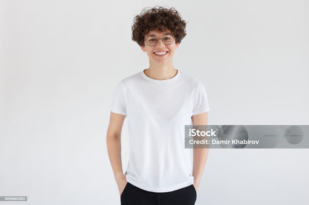 Young european woman standing with hands in pockets, wearing blank white t-shirt with copy space for your logo or text, isolated on gray background Women Stock Photo