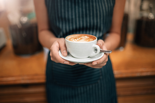 close up of a waitress holding a well poured coffeeCropped Close up of female serving coffee with latte art while standing in coffee shop. Focus on female hands placing a cup of coffee