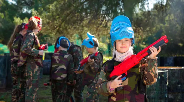 Photo of Teen girl wearing uniform and holding gun ready for playing with friends on paintball outdoor