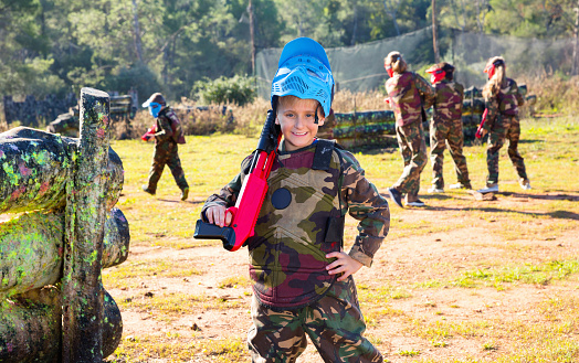 Cheerful positive  boy paintball player in camouflage standing with gun before playing outdoors