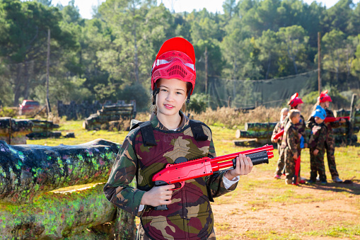 Cheerful positive smiling teen girl wearing uniform and holding gun ready for playing with friends on paintball outdoor