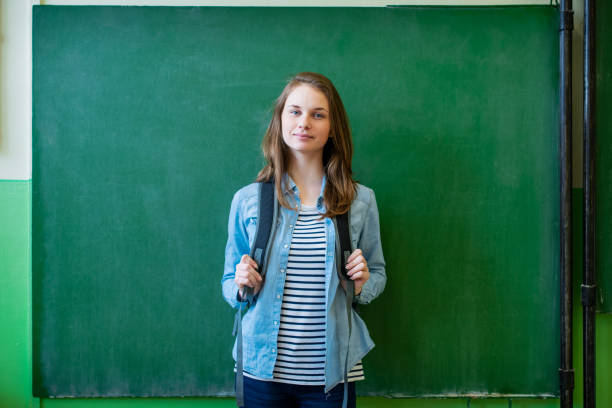 istock Young confident smiling female high school student standing in front of chalkboard in classroom, wearing backpack, looking at camera. Waist up portrait. 1090961884