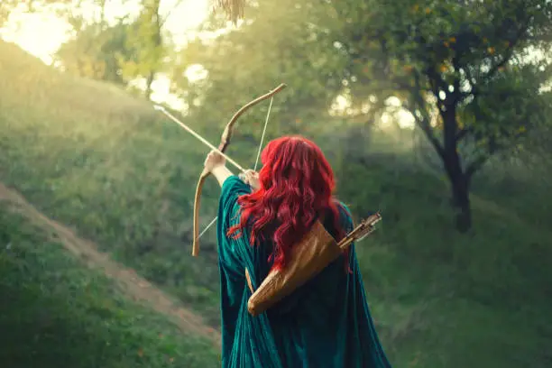 gorgeous huntress pushing her last light to the sun, waiting for salvation during terrible danger, red-haired girl fights for life, emerald velvet cloak, photo without a face from the back.