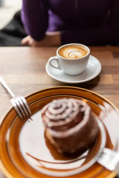 A freshly brewed small flat white coffee in a white cup and white saucer with a brown cinnamon bun on a brown plate and two silver forks out of focus in the foreground.