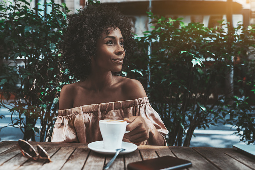 A cheerful young curly female is drinking coffee in a street bar and looking aside; a dazzling happy biracial woman with a cup of tasty hot chocolate is relaxing in a street cafe