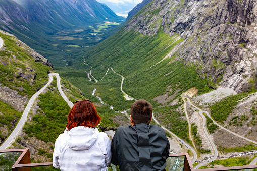 The back view of a red-haired woman and black-haired man is captured on a summer day. The people are looking at Troll Road, Norway in the green valley surrounded by the rocky mountains. The woman is wearing a white jacket, and the man - a black one.