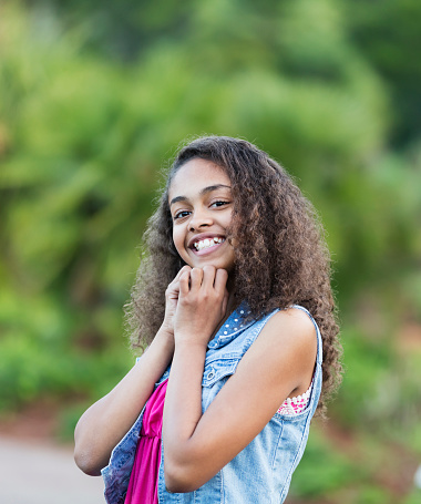 A 13 year old African-American teenage girl standing outdoors in a park, smiling at the camera.