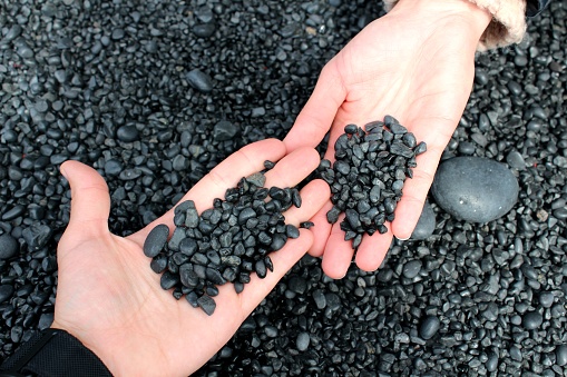 Two travellers visiting Iceland are holding black lava stones from the Reynisfjara beach-sand beach in their hands.