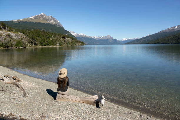 Woman enjoys Acigami Lake Darwin Mountains Tierra del fuego National Park Argentina Sitting next to Acigami Lake, a woman in a hat enjoys the sandy beach, Condor Mountain and the distant Darwin Mountains in Tierra del fuego National Park, Argentina. tierra del fuego national territory stock pictures, royalty-free photos & images