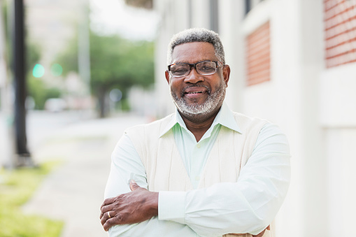 A senior African-American man in his 60s wearing a button down shirt, vest and eyeglasses, looking at the camera with a confident expression.  He is standing outdoors, in the city, arms crossed.