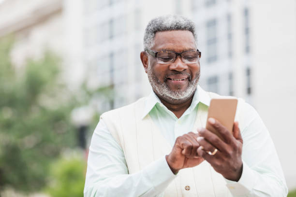 Senior African-American man using mobile phone A senior African-American man in his 60s wearing a button down shirt, vest and eyeglasses, standing outdoors on a city street, looking down at his mobile phone. one senior man only stock pictures, royalty-free photos & images