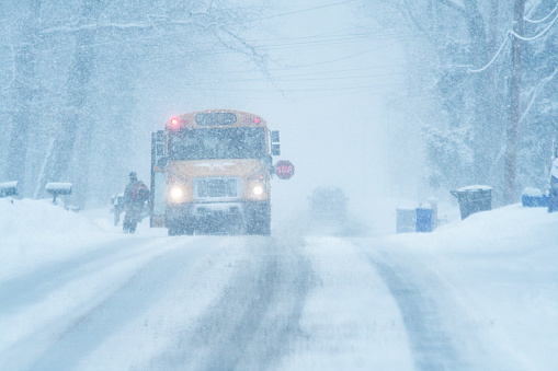A yellow school bus has stopped during a heavy late winter blizzard snow storm to pick up a young elementary school student boy who has just run across the road in front of the bus toward the open door. The bus sits waiting for the unrecognizable child with its emergency warning lights flashing and its illuminated, flashing lights stop sign extended out to stop on-coming traffic. Rural scene in early March near Rochester, NY in western New York State.