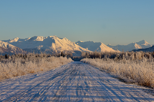 A snow-covered country road leads toward an Alaskan mountain landscape.