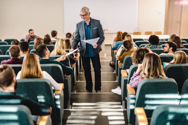 Happy male professor giving his students test results in amphitheater. Happy senior professor talking to his students while giving them test results in lecture hall. amphitheater stock pictures, royalty-free photos & images