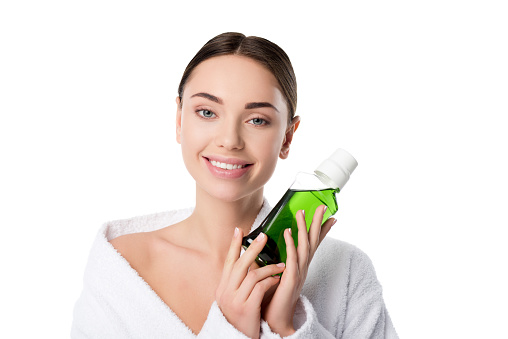beautiful smiling woman in bathrobe holding mouthwash and looking at camera isolated on white