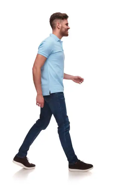 side view of casual man in blue polo shirt walking on white background and smiling, full body picture