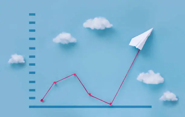 Conceptual origami paper plane pulling business finance growth chart line flying upwards on blue sky background.