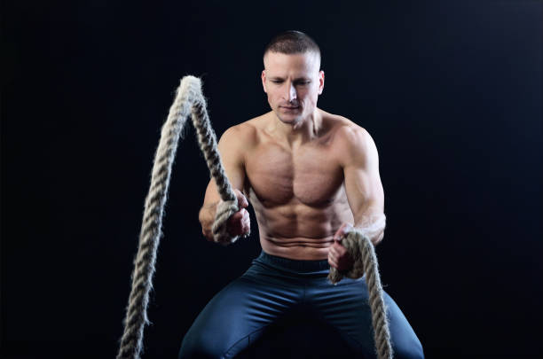 Young muscular man dynamically workout with battle rope on the studio dark background.