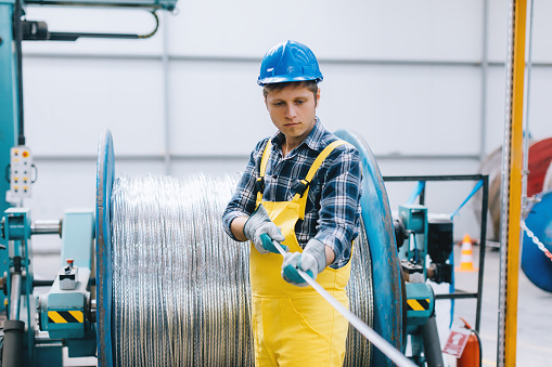 Portrait of a serious technician or industry worker man with crash helmet and worker west making stainless steel cable meter wire roll and working in a large  factory warehouse. XXXL size taken with Canon 5D Mark4