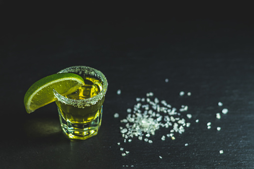 Mexican Gold Tequila shot with lime and salt on black stone table surface, selective focus, shallow depth of the field, copy space.