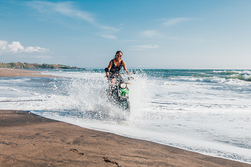 Young afro american woman riding enduro motorbike on the beach coastline splashing the water