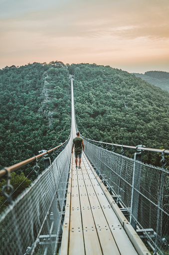 Photo of young Man walking on a hanging bridge in Germany during the summer day, Geierlay, The longest pedestrian bridge in Germany, suspension swing bridge. Young Man standing alone outdoor and admiring view Travel Lifestyle concept with rope bridge background and sunset.
