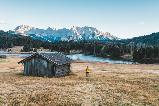 Photo of a man looking at scenic view of Gerald lake in Alps while standing next to the wooden house during autumn day. Lonely male traveler in wintertime at Lake Gerald enjoying the view.