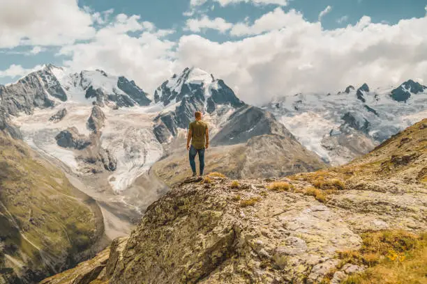 Young man enjoying the stunning summer view of Corvatsch peak glacier from St Moritz in Switzerland. Photo of  young Traveler Man relaxing meditation with serene view mountains landscape Travel Lifestyle hiking concept summer vacations outdoor. Day dreamer,human strength and psychology concept.