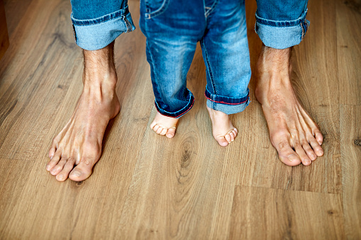 Low section of father and son standing on hardwood floor. High angle view of man is with baby boy at home. They are barefoot.