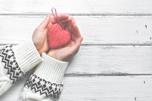 Red heart in the hands of a young woman in a white wool sweater with an ornament on a white wooden rustic background. Valentine's Day. Top view, place for text.