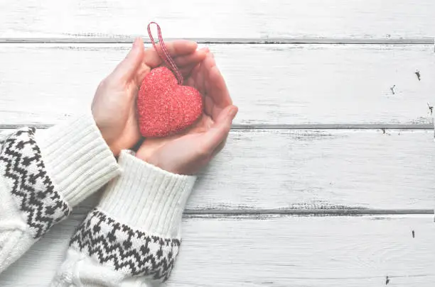 Photo of Red heart in the hands of a young woman in a white wool sweater with an ornament on a white wooden rustic background. Valentine's Day. Top view, place for text.