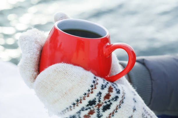 ウールのミトンで手は、熱いお茶やコーヒーと赤いマグカップを保持します。川や海の雪に覆われた銀行に冷ややかな日当たりの良い日にピクニック。冬の散歩に温暖化のコンセプト - glove winter wool touching ストックフォトと画像