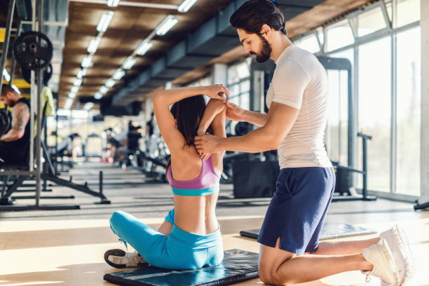 serious bearded personal trainer helping woman to stretch arms. woman sitting on the mat, gym interior. - secrecy instructor exercising individuality imagens e fotografias de stock